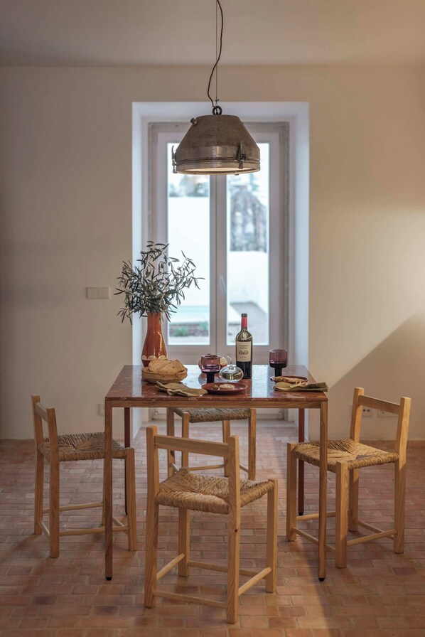 Un comedor con mesa de madera, sillas de mimbre, lámpara colgante y ventana con vistas.