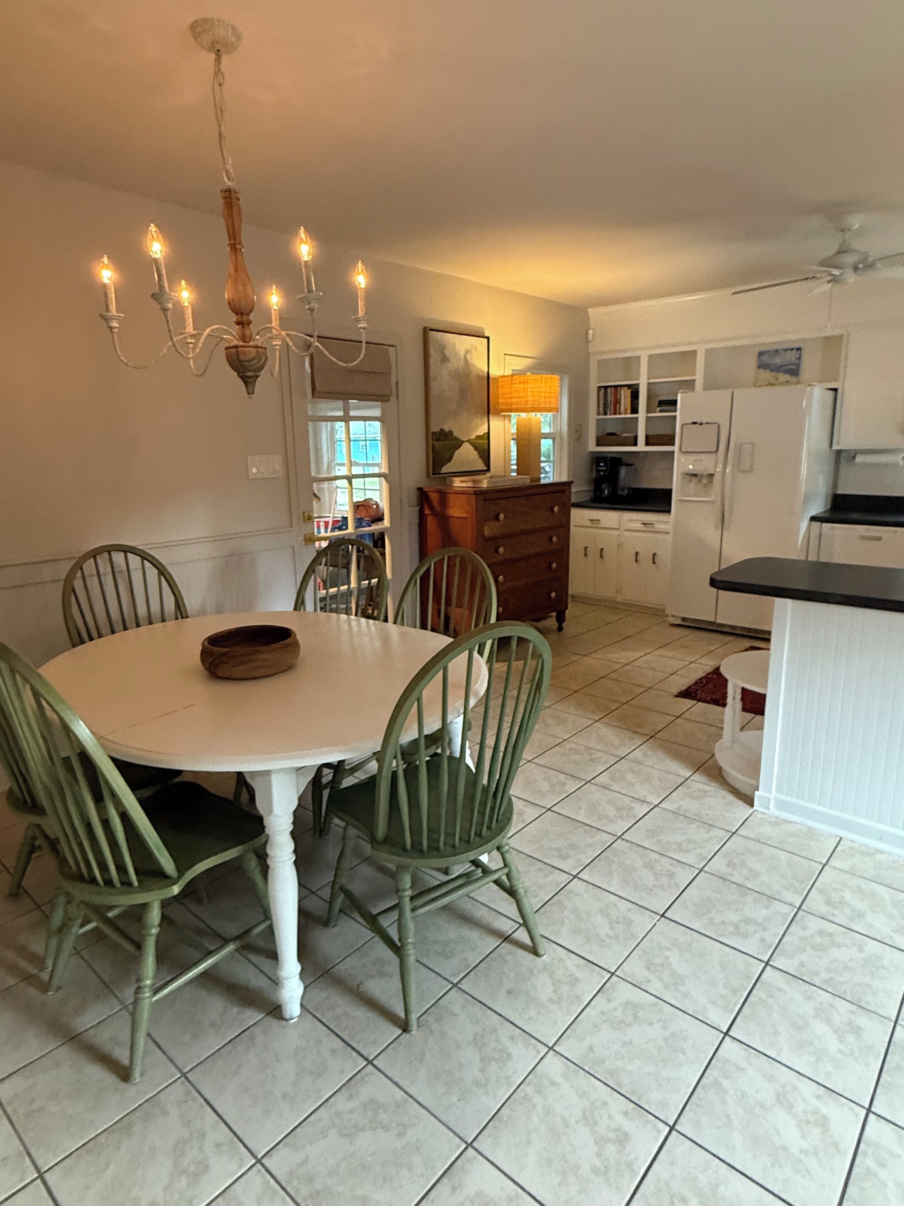 A dining area with a round table, four chairs, a chandelier, and a kitchen in the background.