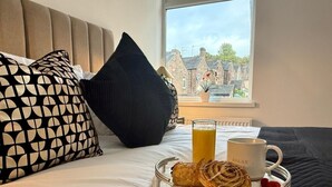 Un lit bien fait, avec un plateau de petit-déjeuner comprenant des viennoiseries, un verre de jus d’orange et une tasse, placé devant une fenêtre donnant sur des immeubles.