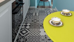 Une petite cuisine équipée d’un évier, d’ustensiles et d’une chaise. On y trouve une table avec deux bols. Le sol est recouvert de carreaux décorés.