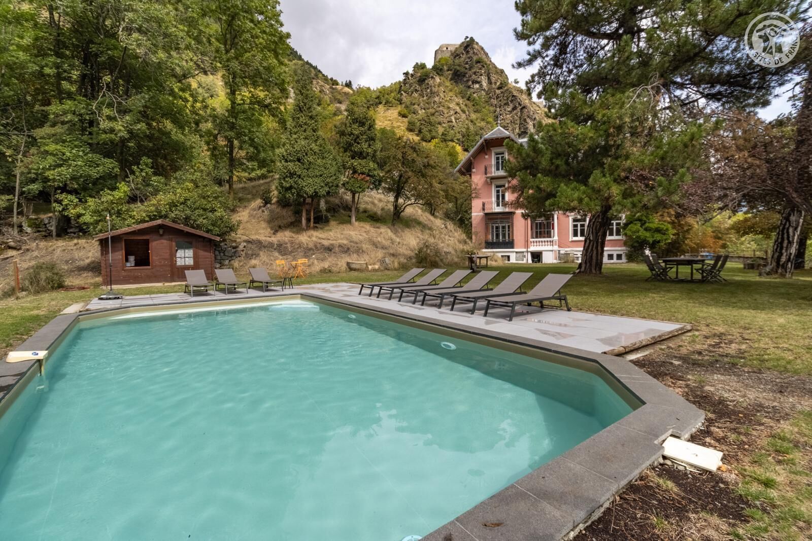 Une piscine avec des chaises longues et un petit chalet en bois, entourés d’arbres et avec un grand bâtiment en arrière-plan.
