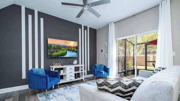 Un salón moderno con televisor de pantalla plana, dos sillones azules, un sofá blanco, un ventilador de techo y un amplio ventanal corredera al exterior.