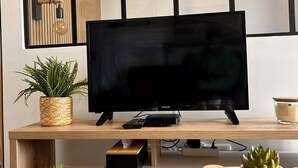 Una sala de estar moderna con un televisor de pantalla plana sobre un soporte de madera, una maceta tejida, una planta pequeña en maceta, una mesa de madera redonda con frascos, y una lámpara de pared.