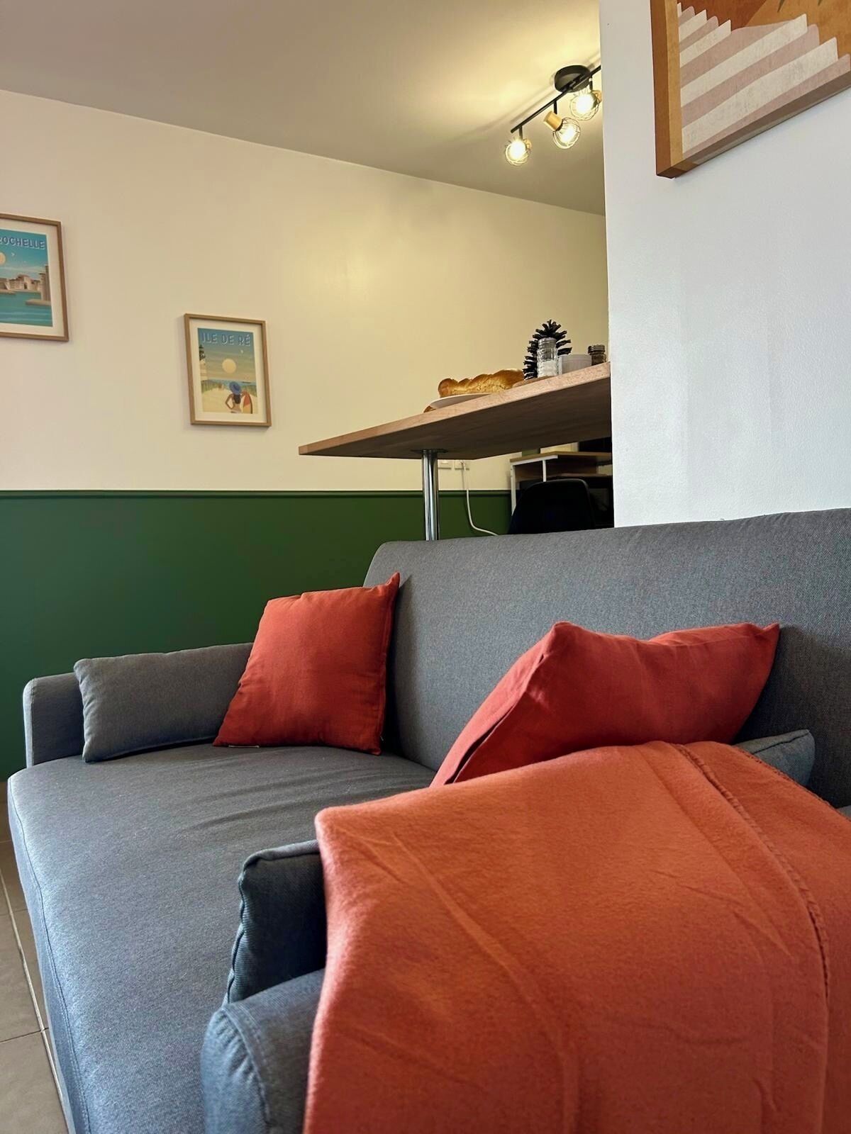 A modern living room with a grey sofa and red cushions, a wooden table with a lamp, and two framed pictures on the wall.