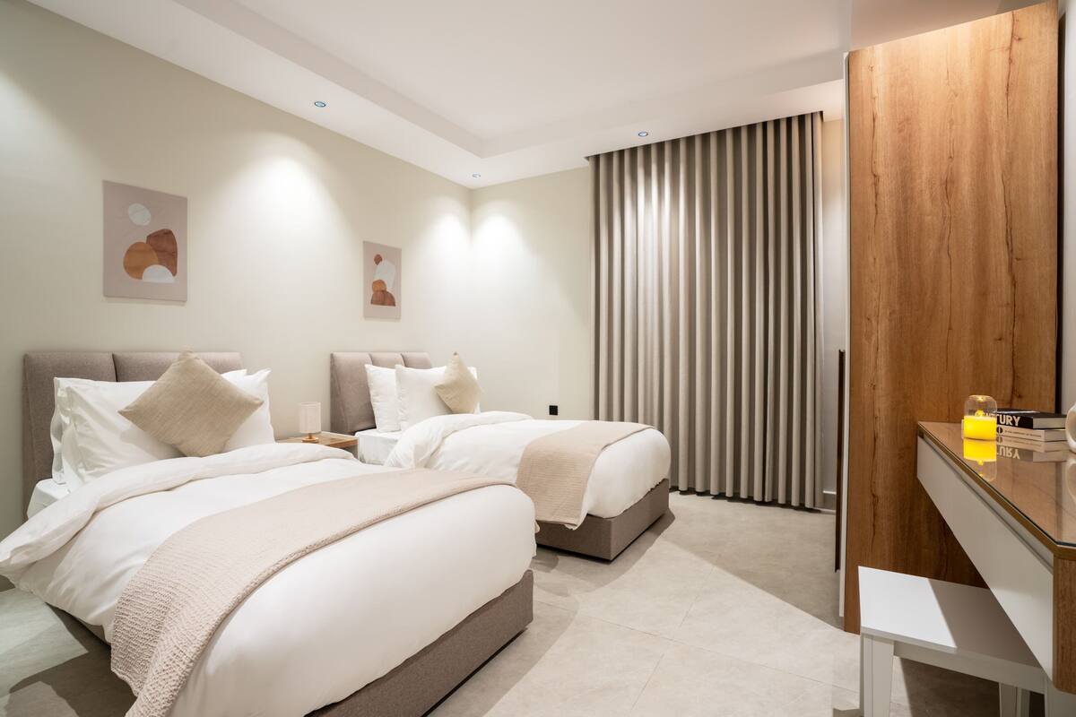 Quarto de hotel moderno com duas camas, um guarda-roupa de madeira, uma mesa de cabeceira com abajur e uma janela grande com cortinas.