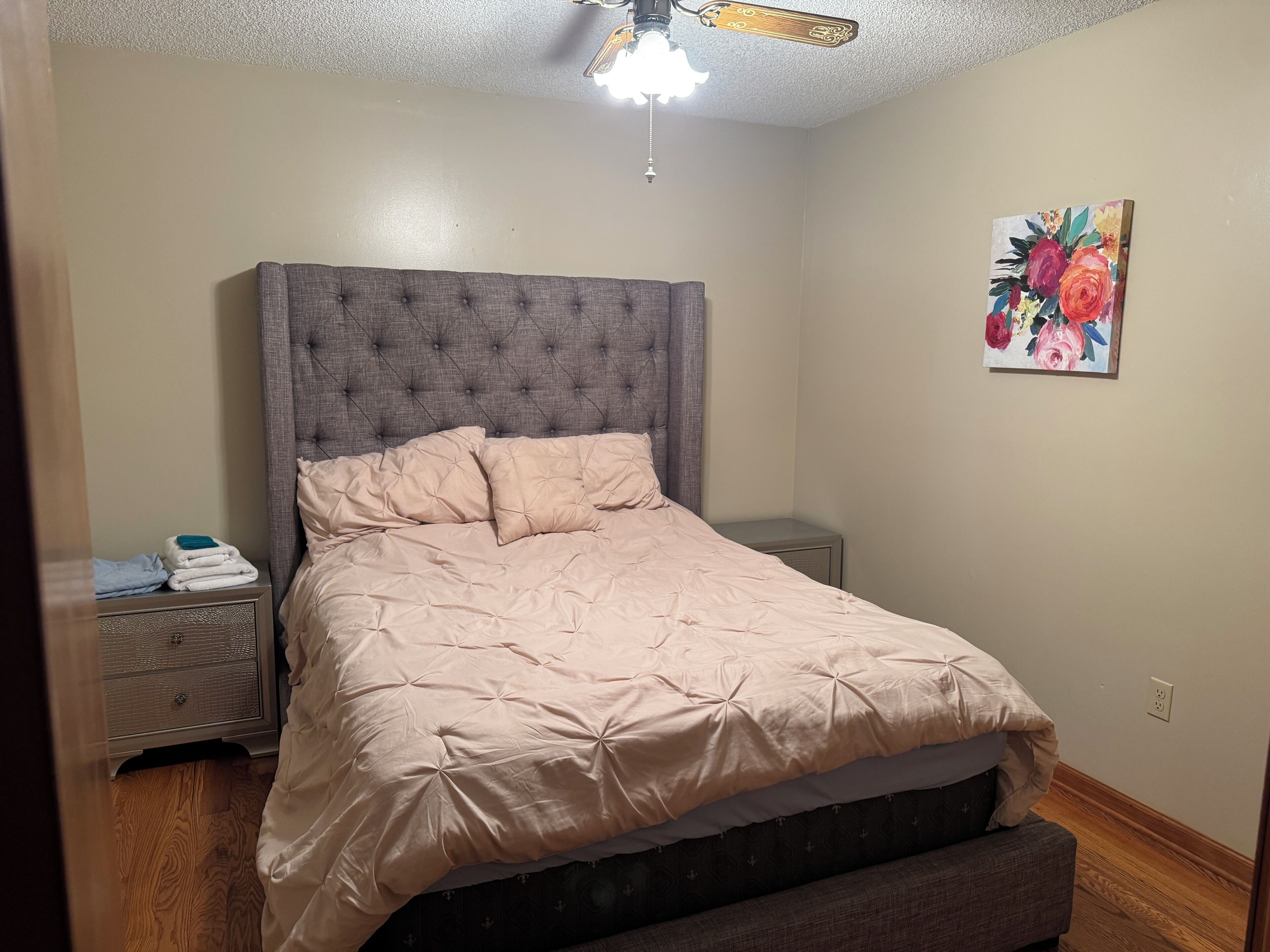Un dormitorio con cabecera capitonada, cama con colcha color beige, mesita de noche con lámpara y una obra de arte floral en la pared.