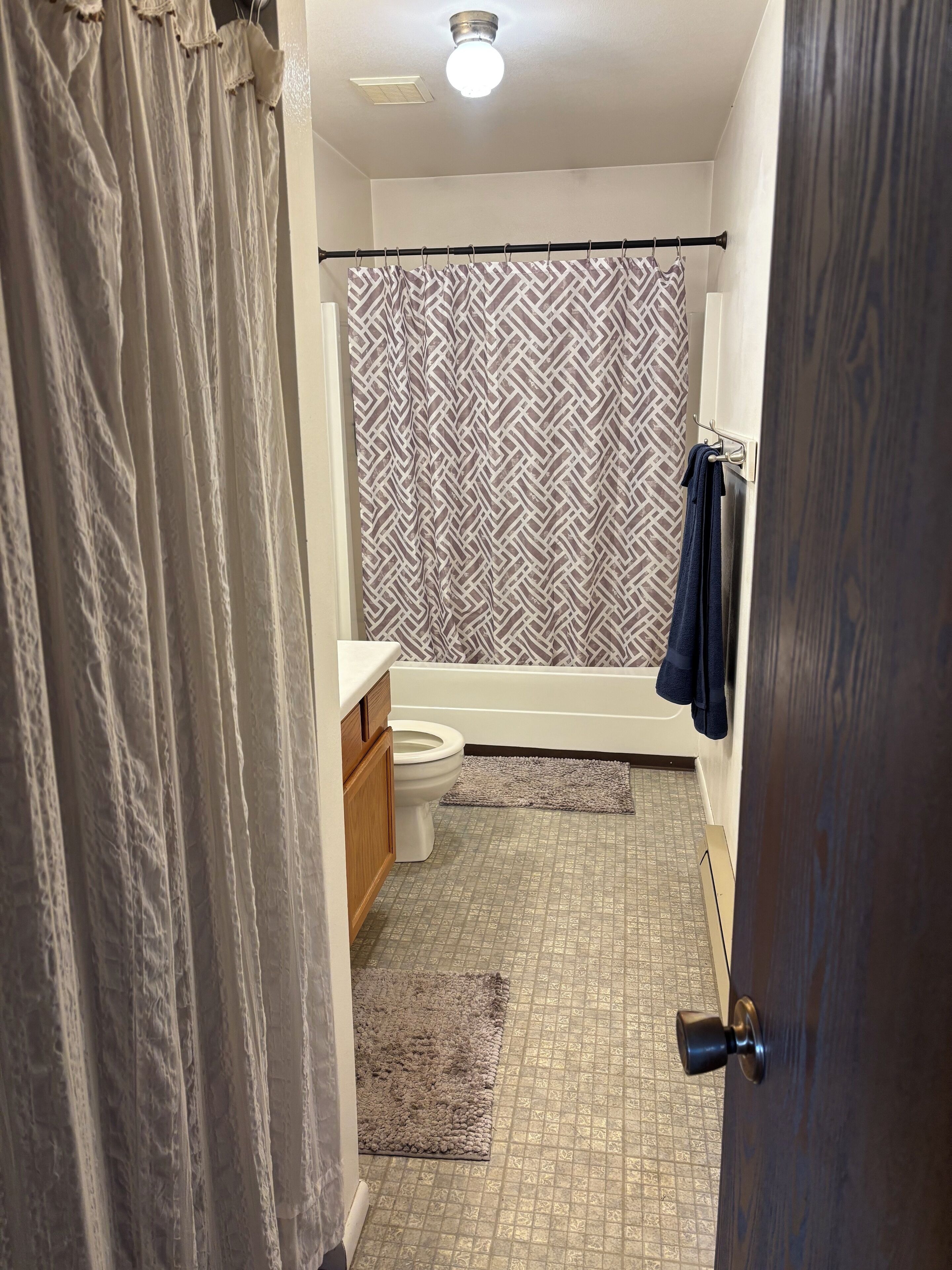 A bathroom with a toilet, a bathtub, a shower curtain with a geometric pattern, a towel rack, and a patterned floor.