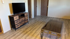 A room with a wooden floor, a TV on a wooden cabinet, and a glass coffee table with metal legs. - Charming 2-bedroom Getaway in Broadus (Broadus)