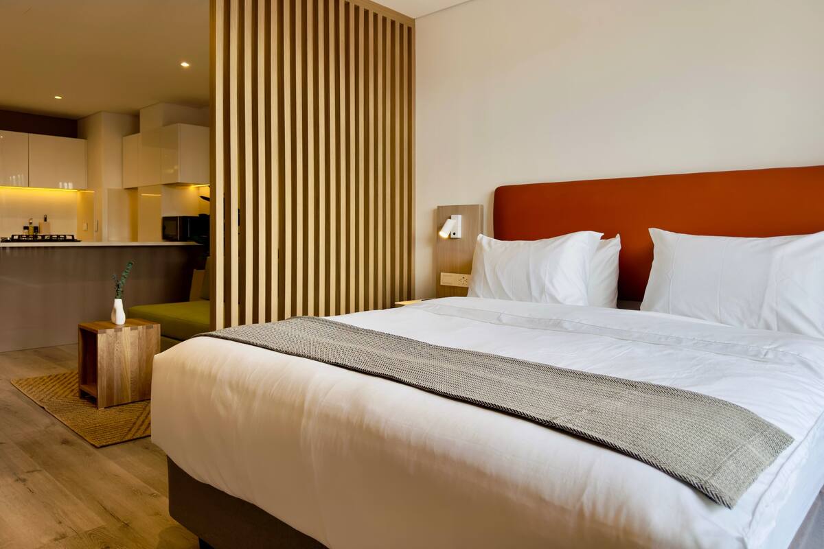 A modern hotel room with a large bed, a wooden partition, a kitchenette, and a small side table with a vase.