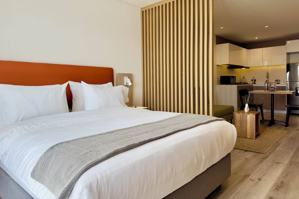 A modern hotel room with a large bed, a wooden partition, a small kitchen area, and a dining table.