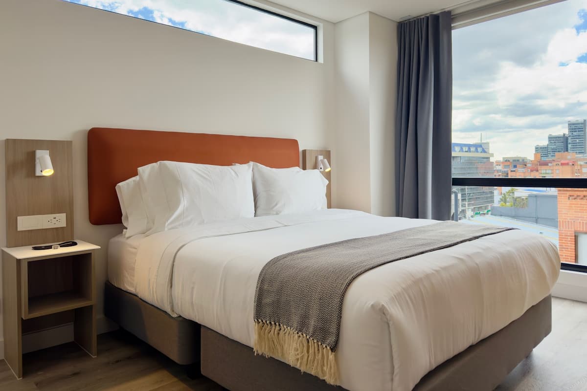 A modern hotel room with a large bed, a nightstand, a wall-mounted lamp, and a view of the city through a large window.