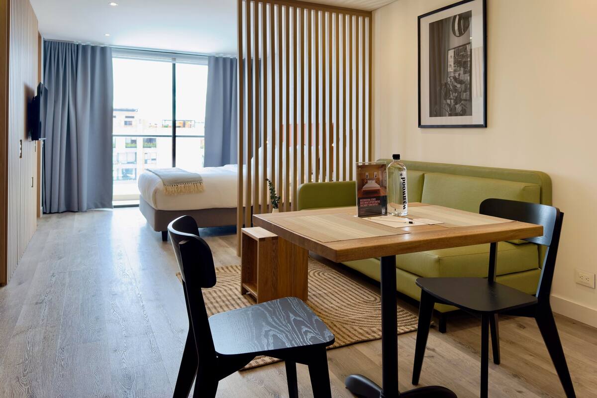 A modern hotel room with a bed, a green sofa, a wooden dining table, and black chairs.