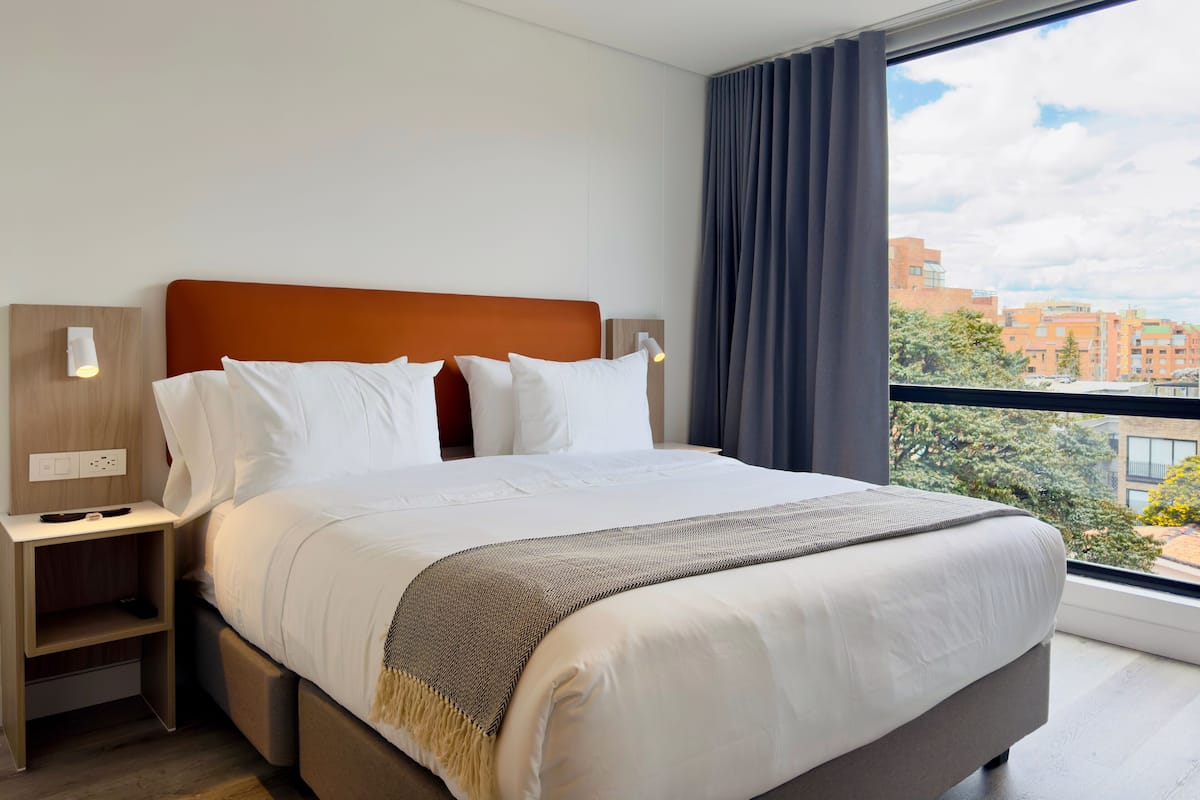 A modern hotel room with a large bed, two bedside tables, a nightstand, and a view of the city through large windows.