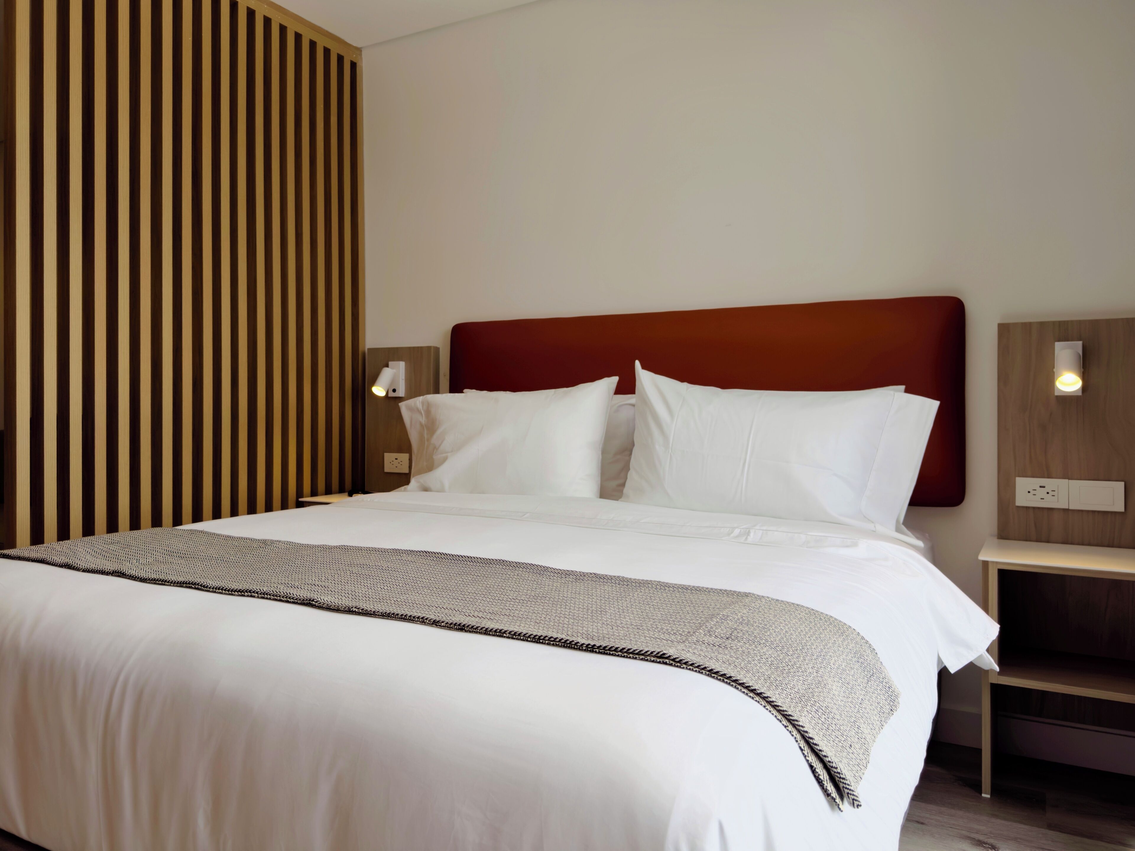A hotel room with a large bed, a wooden headboard, two bedside tables with lamps, and a wall-mounted light fixture.