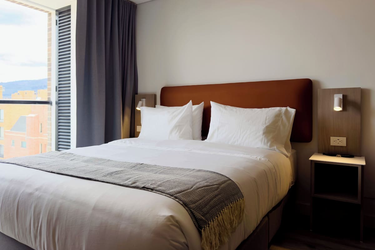 A hotel room with a large bed, a nightstand, a wall-mounted light, and a view of buildings through a window with curtains.