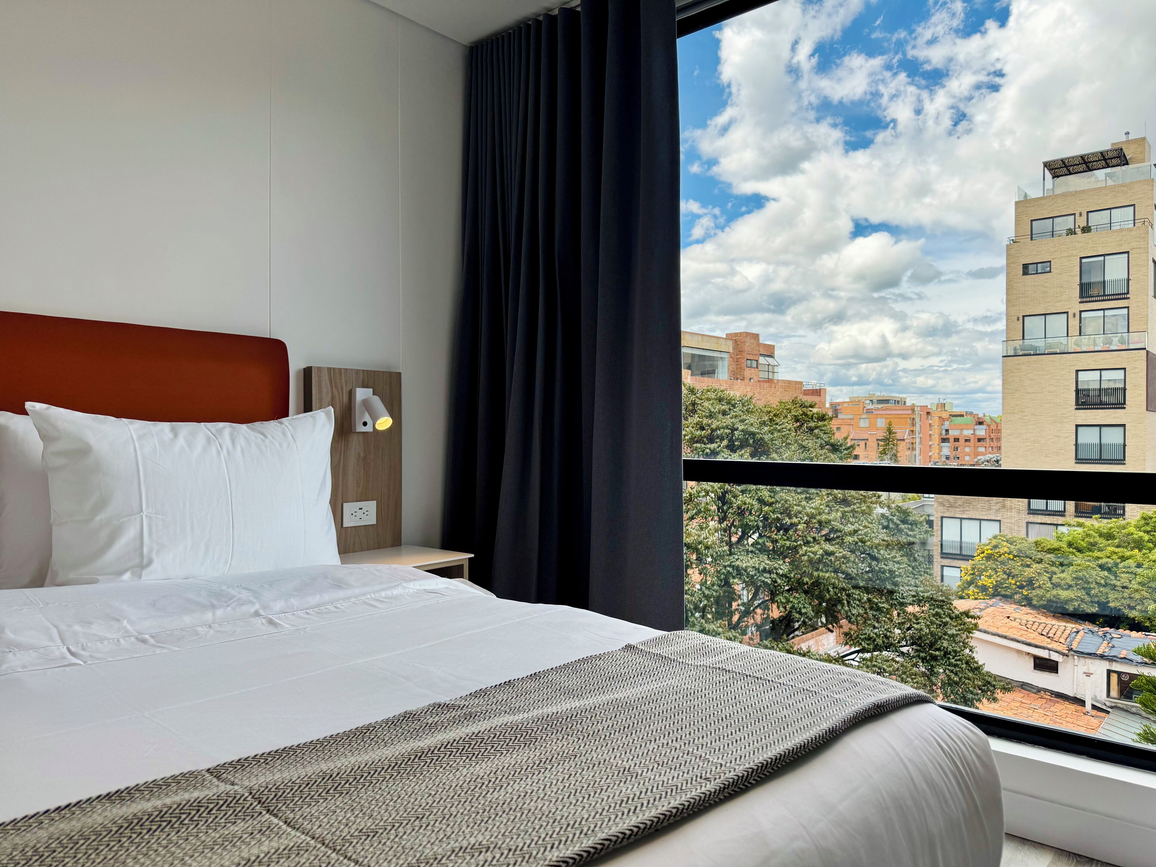 A hotel room with a bed, a nightstand, a wall-mounted lamp, and a window with a city view.