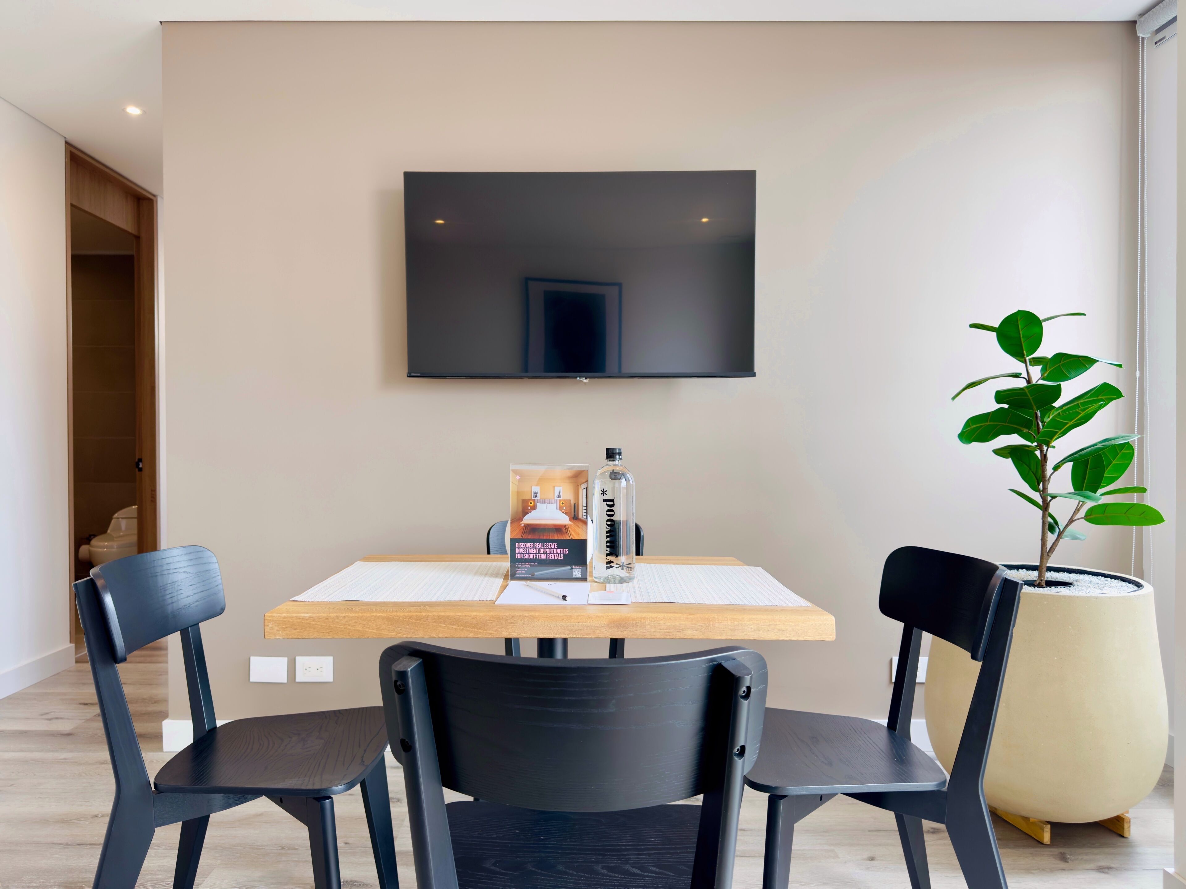 A modern dining area with a table, chairs, a potted plant, and a flat-screen TV mounted on the wall.