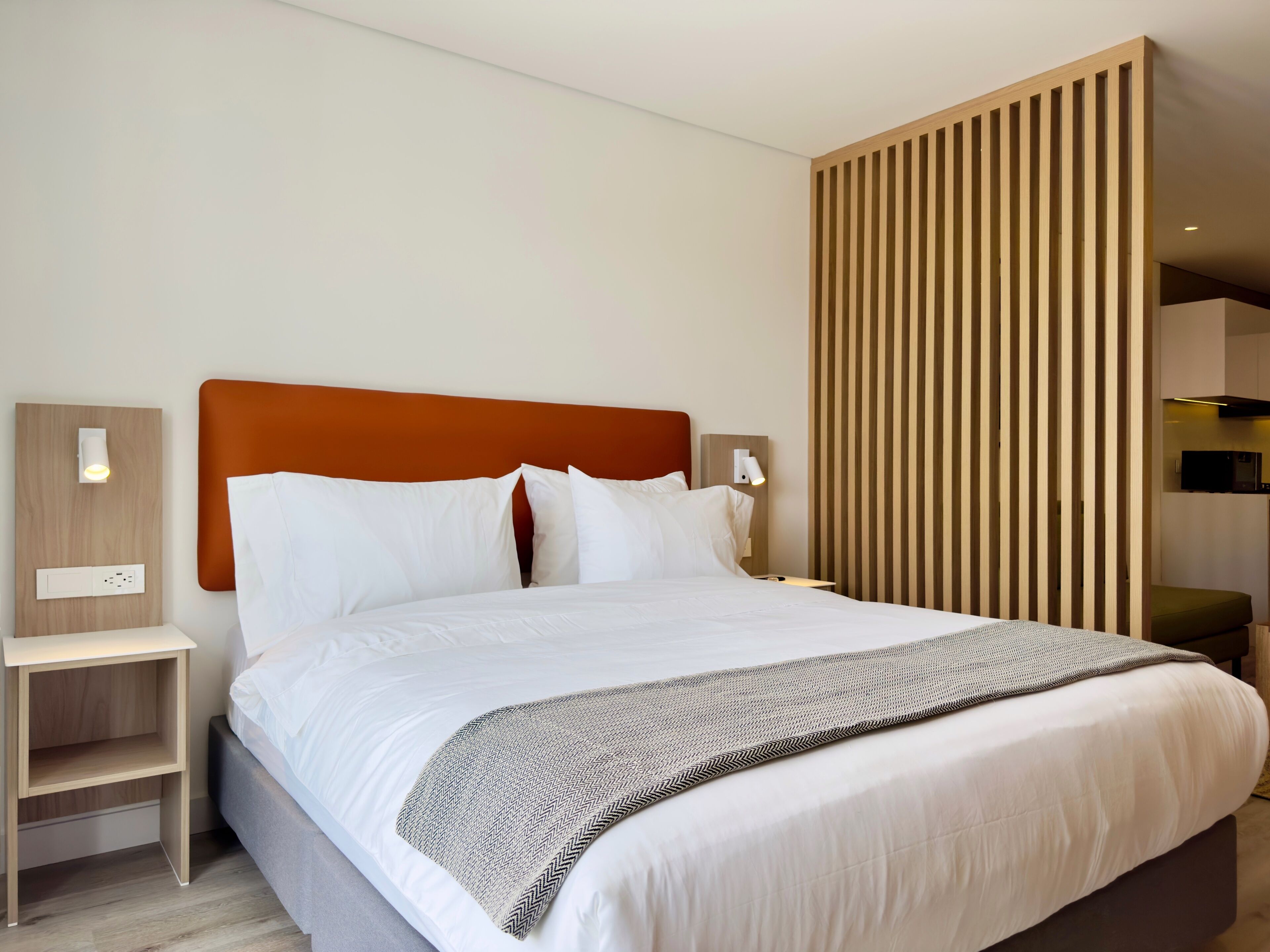 A modern hotel room with a large bed, wooden headboard, bedside tables, and a built-in TV.