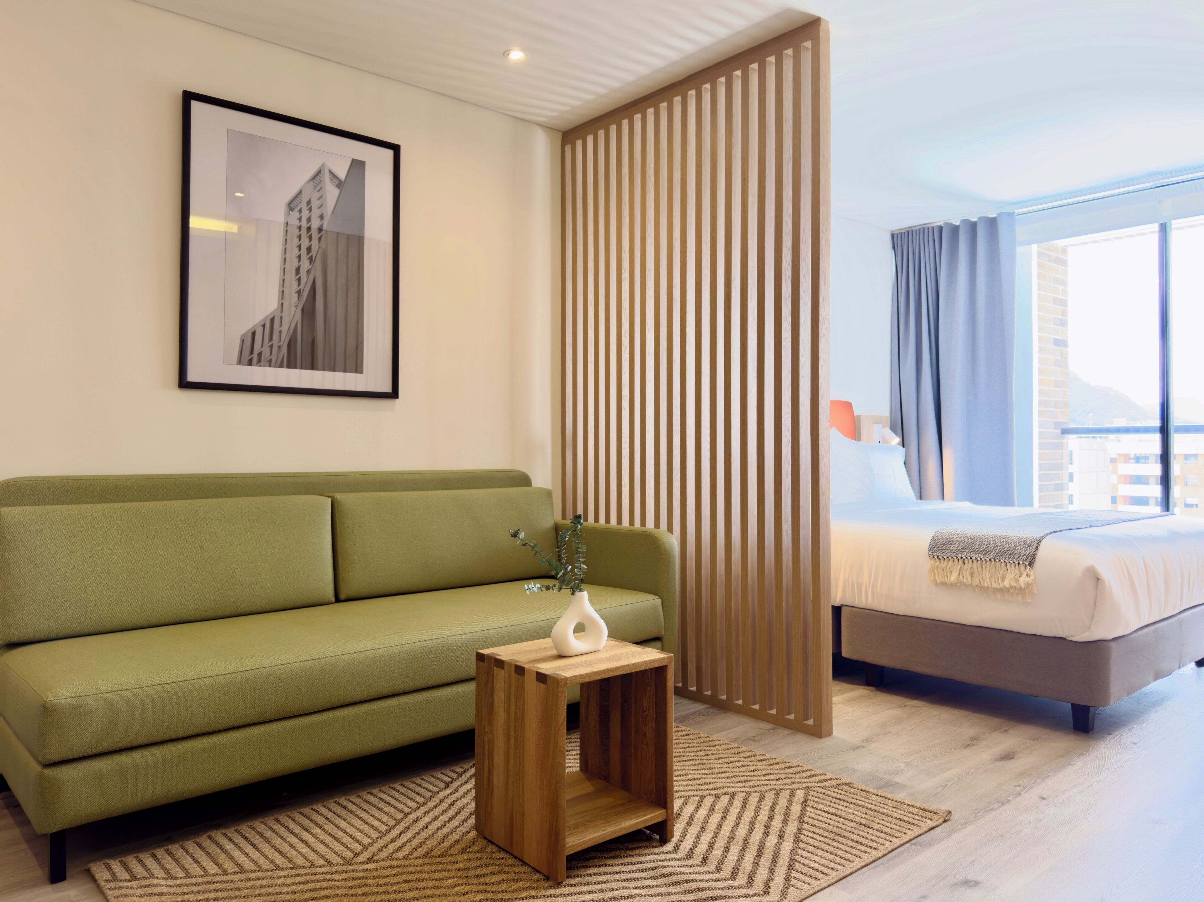 Une chambre d’hôtel moderne avec un canapé vert, une table d’appoint en bois surmontée d’un vase, une grande fenêtre avec des rideaux et un tableau encadré accroché au mur.