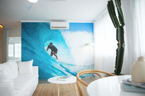 Un salon avec une grande fresque représentant un surfeur sur une vague, un canapé blanc, une table ronde et un cactus.