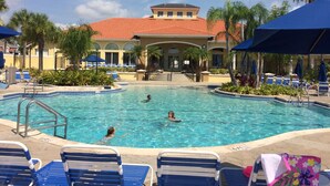 Ein Swimmingpool mit Liegestühlen und Sonnenschirmen, umgeben von Palmen und einem Gebäude mit rotem Dach.