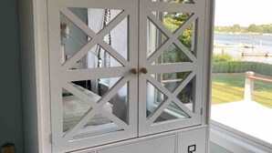 A white cabinet with glass doors and drawers, located next to a window with a view of a dock and greenery.