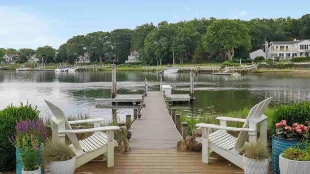 A wooden dock leading to a calm lake with boats, surrounded by greenery and houses.