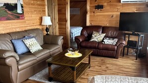 Un salon chaleureux aux murs et au plancher en bois, avec un canapé en cuir marron, un fauteuil marron, une table basse et un meuble pour la télévision, sur lequel est posée une télévision à écran plat.