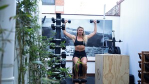 A person exercising with dumbbells in a room with a large wooden cabinet and a wall-mounted rack of dumbbells.