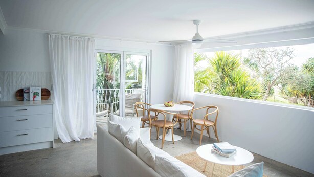 A bright, airy room with a white sofa, a dining table with chairs, and a balcony with a view of greenery.