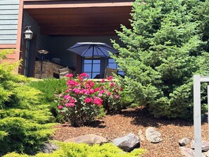 A building with a covered entrance, a patio area with a table and chairs, and a garden with pink flowers and green shrubs.