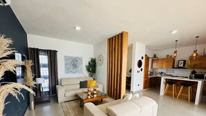 A modern living room with a sofa, coffee table, and decorative plants. There is a kitchen area with wooden cabinets and bar stools.