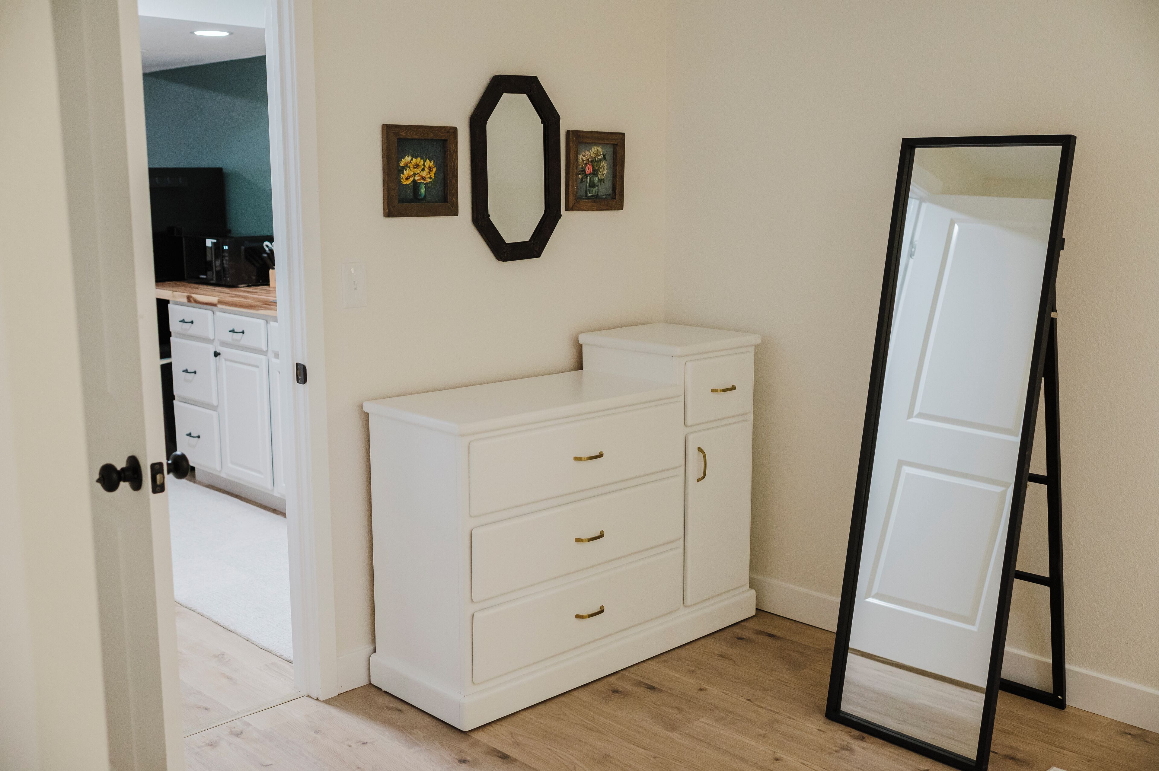 Une pièce avec une commode blanche, un miroir et une porte qui donne sur la cuisine.