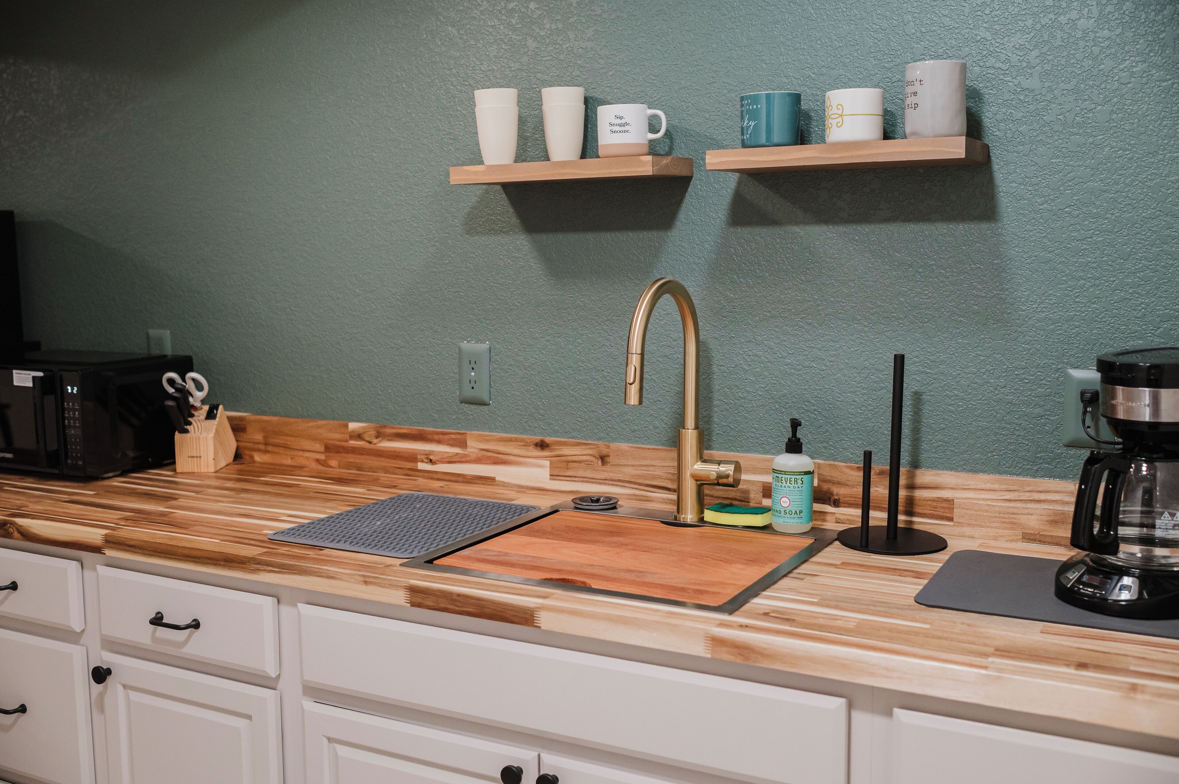 A kitchen with a wooden countertop, a stainless steel sink, a coffee maker, and a shelf with cups.