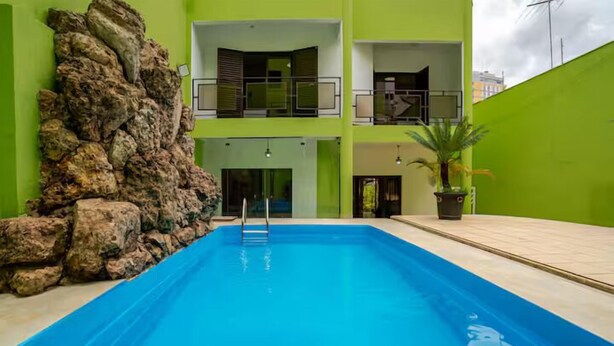 A swimming pool with a rock wall on one side, a green building with balconies, and a potted plant. - Casa Liberdade (São Paulo)