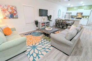 Una sala moderna con un sofá gris, una mesa de centro y un tapete colorido. Se ve, al fondo, una zona de comedor con una mesa y sillas.