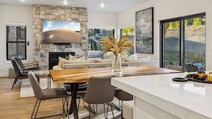 A modern living room with a stone fireplace, a large flat-screen TV, a dining area with a wooden table, and a view of a natural landscape through large windows. - Hot Tub, BBQ & Mtn Views Near Deer Valley & Park City Deer Valley East Retreat by Avantstay (Heber City)