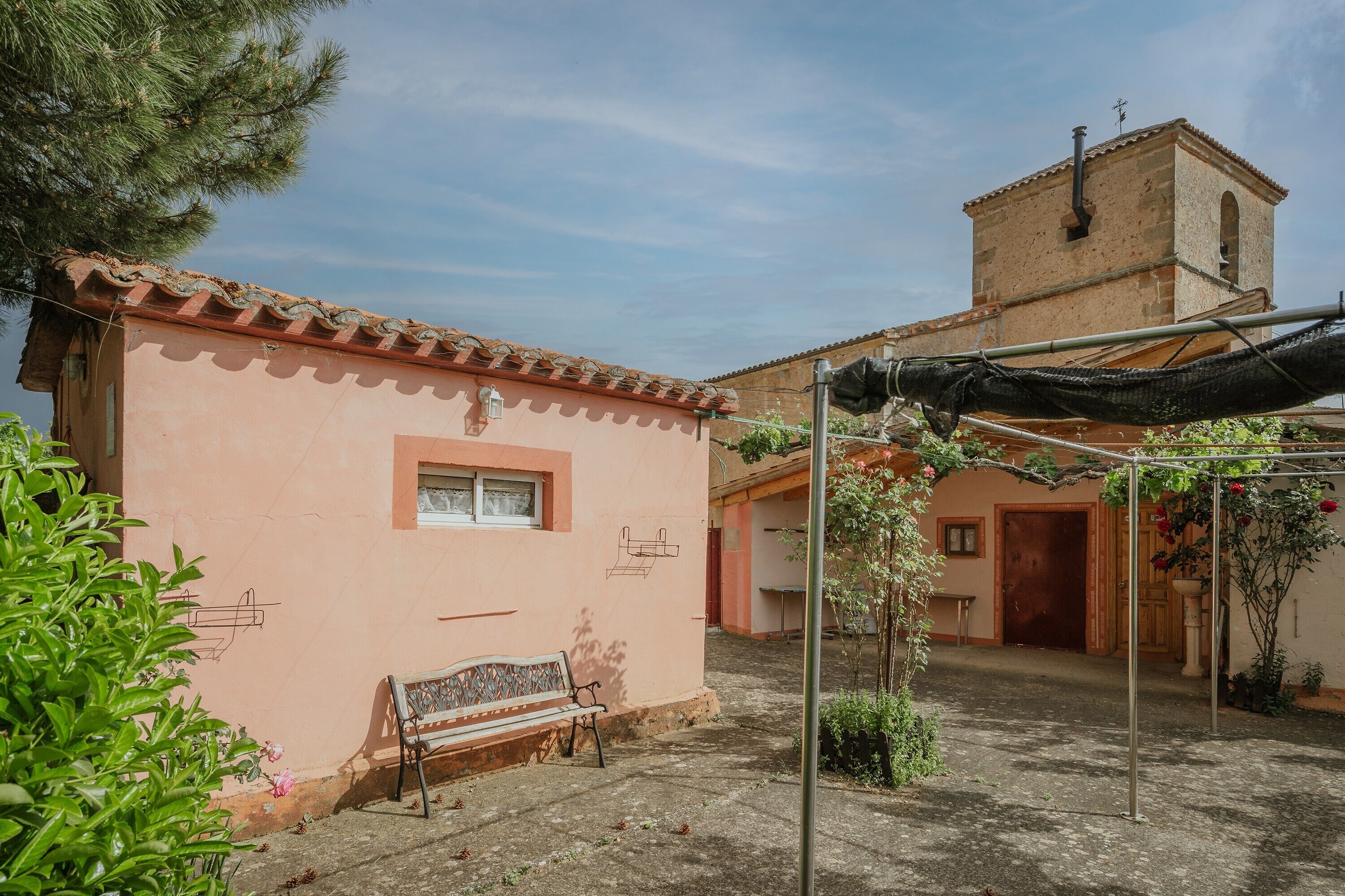 A courtyard with a bench, a pergola with climbing plants, a building with a tiled roof, and a bell tower.