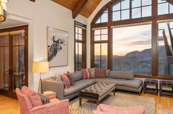 A spacious living room with a large sectional sofa, a small ottoman, and a floor lamp. There is a large window with a view of mountains and a framed artwork on the wall.