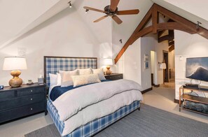 A bedroom with a large bed, wooden beams, a ceiling fan, and a TV on a stand.