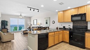 A modern kitchen with granite countertops, wooden cabinets, and black appliances. There is a dining area with a table and chairs, and a balcony with a view of the ocean. - 2 Beachfront Condos BBQ Balconies, Resort Pool, Hot Tub + Direct Beach Access (Panama City Beach)