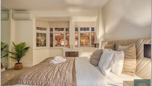 A bedroom with a large bed, a potted plant, and a view of a lit building through the windows.