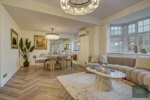 Un salon spacieux agrémenté d’un grand lustre, d’un canapé, d’un coin repas et offrant une vue dégagée grâce à de grandes fenêtres.