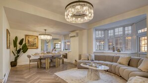 A spacious living room with a large window, a chandelier, and a dining area with a wooden herringbone floor.