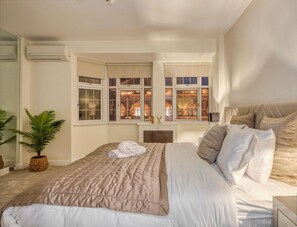 Une chambre à coucher avec un grand lit, une plante en pot et une vue sur un bâtiment illuminé, visible à travers les fenêtres.
