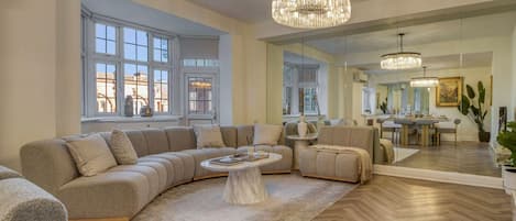 A spacious living room with a large sectional sofa, a round coffee table, and a chandelier. There is a dining area visible through a glass partition.