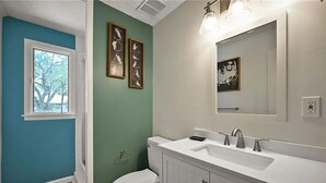 Une salle de bain avec un lavabo blanc, un miroir et des toilettes. Deux tableaux encadrés ornent le mur, et une fenêtre offre une vue sur les arbres.