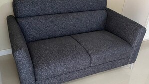 A grey upholstered sofa with a metallic frame, positioned against a plain wall with a tiled floor.