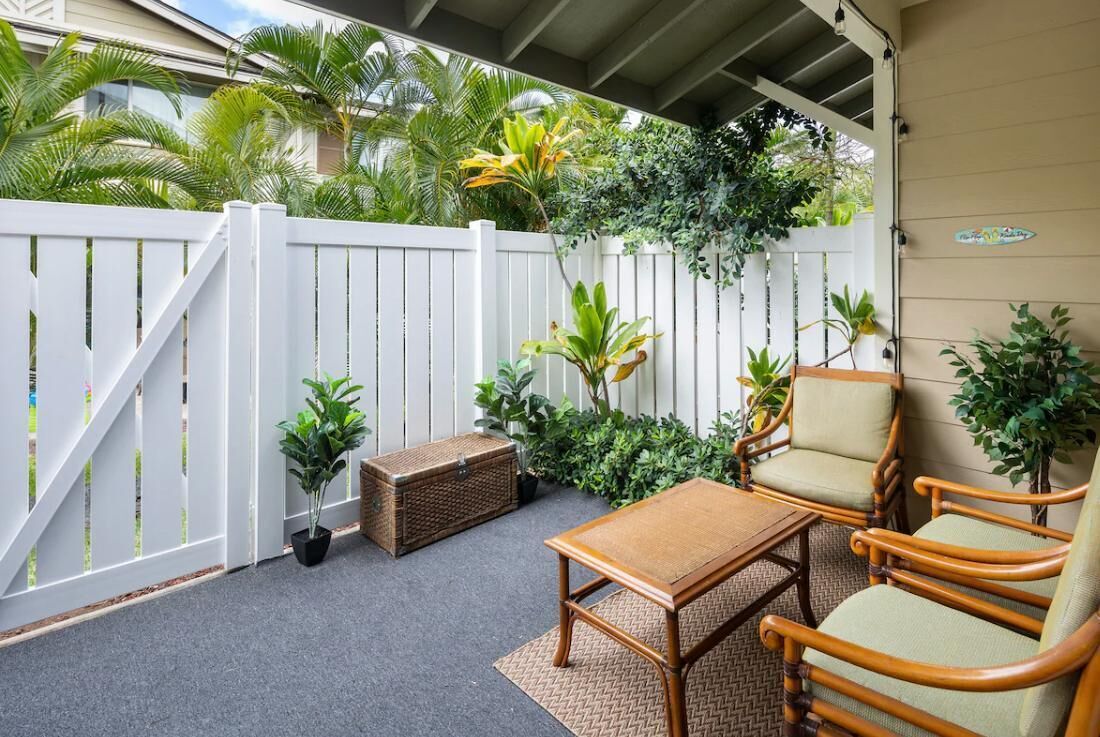 A covered patio with a white fence, wicker furniture, and tropical plants.