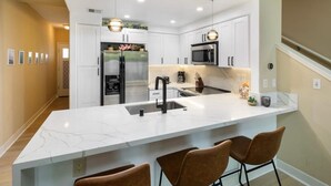A modern kitchen with white cabinetry, a large island with bar stools, stainless steel appliances, and pendant lighting. - Modern Family Beach Villa near shops and dining (Kapolei)
